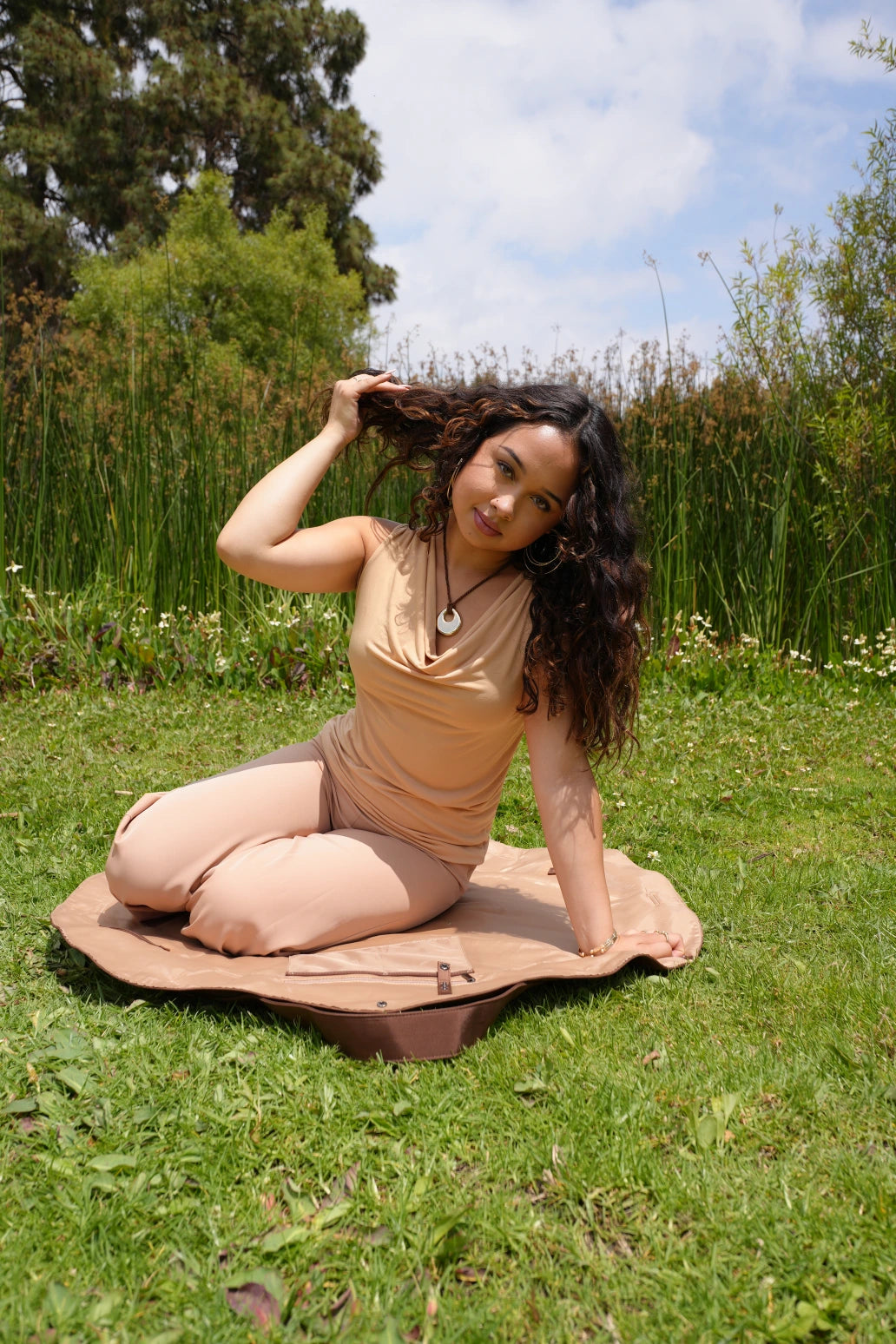 Woman sitting on a mat outdoors with greenery and blue sky in the background
